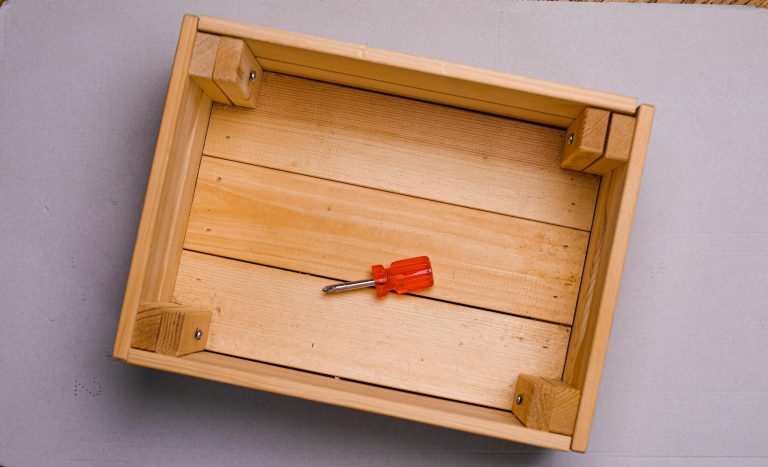 Flat lay of a red screwdriver inside an empty wooden box on a gray background.